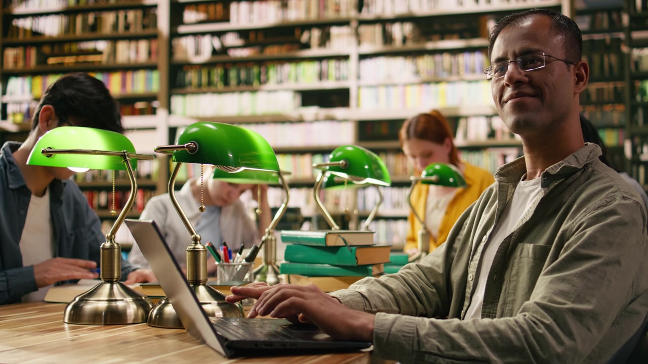 Portrait of indian smiling tutor looking up from laptop screen at library desk