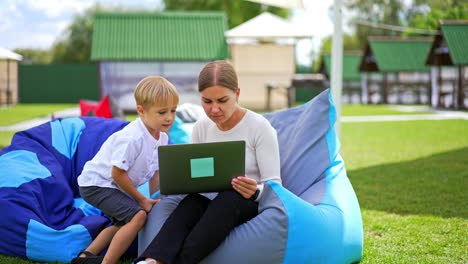 blonde woman with laptop in her hands sits in bean bag chair. mother showing something on the screen to her six-year old son. blurred backdrop.