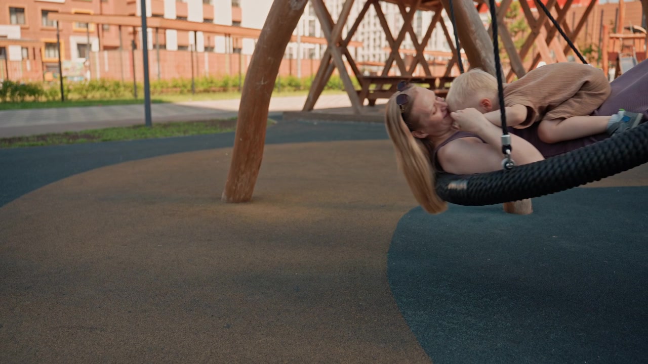 Una donna e un bambino piccolo condividono un'altalena in un parco giochi all'aperto. La scena cattura la loro interazione mentre si dondolano dolcemente con uno sfondo che presenta strutture di gioco in legno e un'area residenziale.