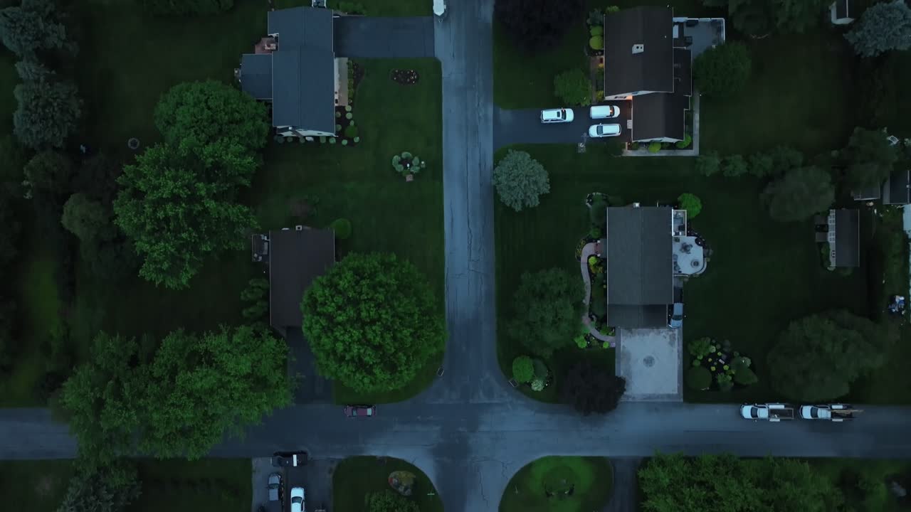 Straight street with single family houses in green suburbia of town in America. Aerial top down flyover shot. Green grass in yards and garden. Roofing shot of one family homes in Usa. Dusk scene