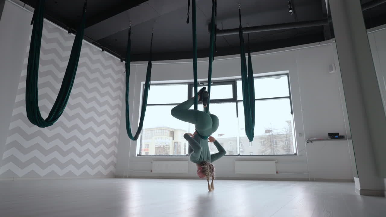 joven mujer hermosa practicando en un columpio de estiramiento aéreo. mujer joven gira su cabeza boca abajo en una hamaca para el yoga aéreo. mujer bastante madura enfocada haciendo yoga aéreo en el gimnasio