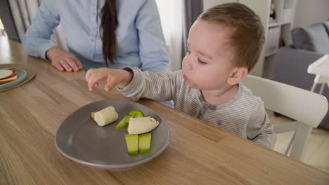 lindo bebé jugando con comida mientras se sienta a la mesa con su madre en la sala de estar