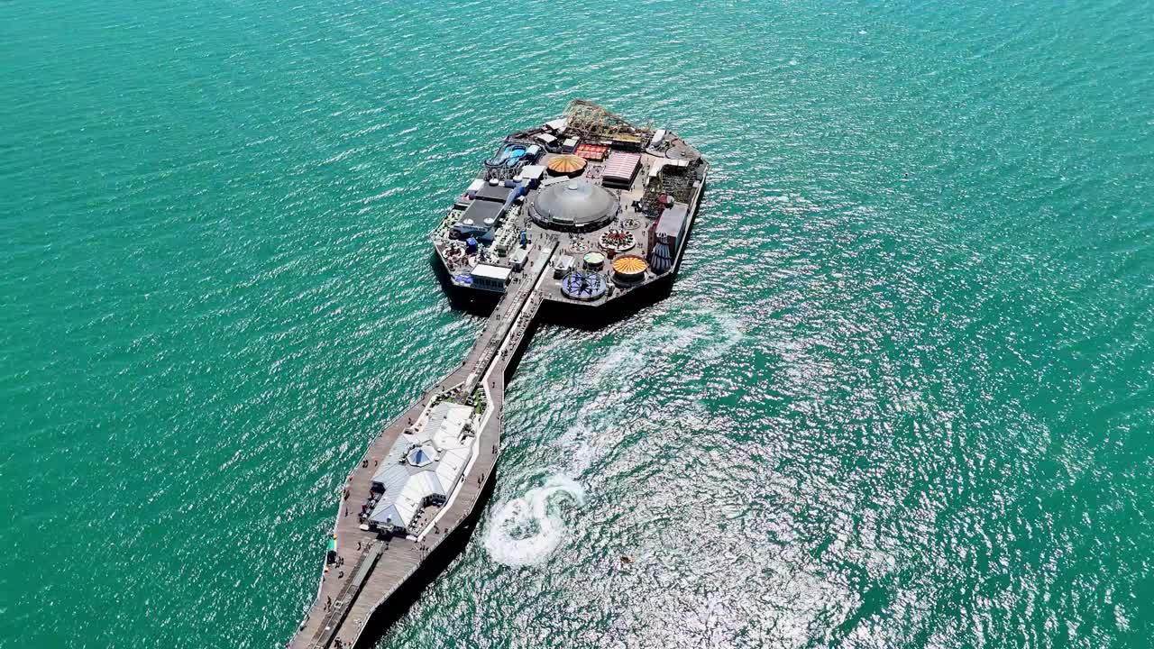 Aerial summer view of Brighton's iconic Palace Pier, Sussex, UK. Sparkling sea, boats and jet skis circling below in the sun. Capturing vibrant coastal life from above.