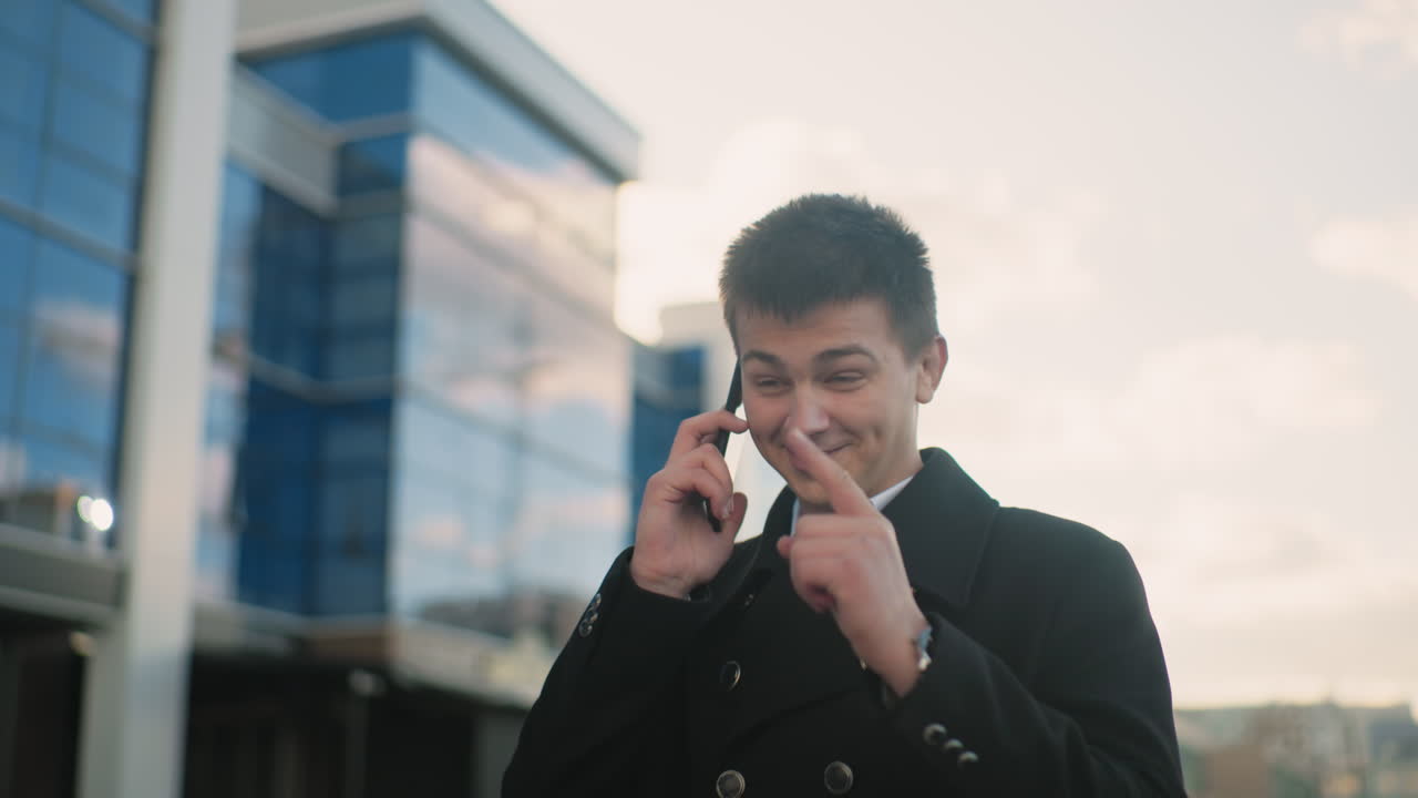 흑색 코트를 입은 사업가가 행복하게 전화를 하고 있으며, 현대적인 유리 건물, 부드러운 빛, 그리고 활기찬 야외 풍경이 있는 도시 지역에서 흥분하고 있으며, 긍정과 직업적 성공을 전달합니다.