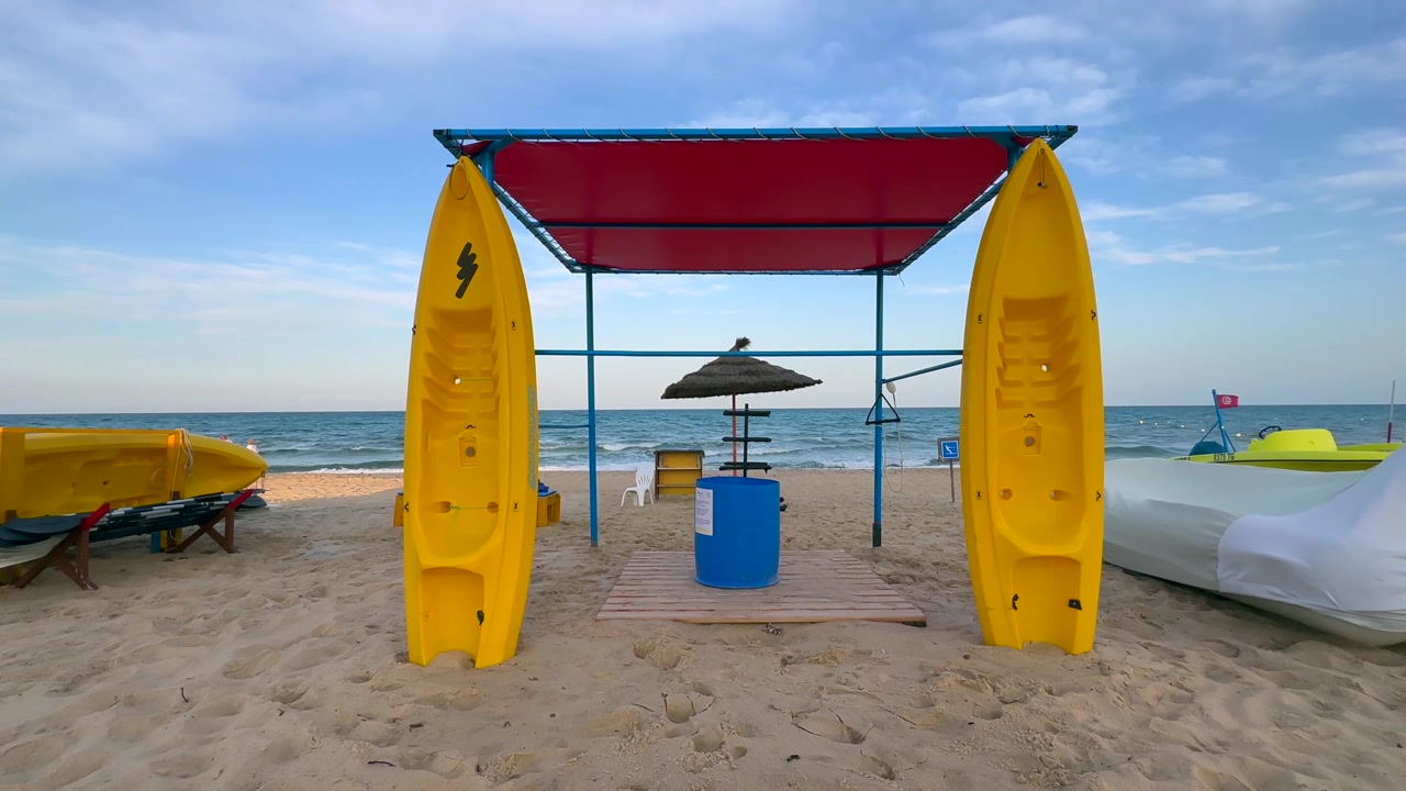 Due kayak giallo brillante sono posizionati verticalmente accanto a un semplice chiosco per il noleggio in spiaggia di fronte a un mare calmo, mentre una coppia in camicia bianca passeggia lungo la spiaggia in un angolo romantico baciato dal sole. Hammamet, Tunisia.