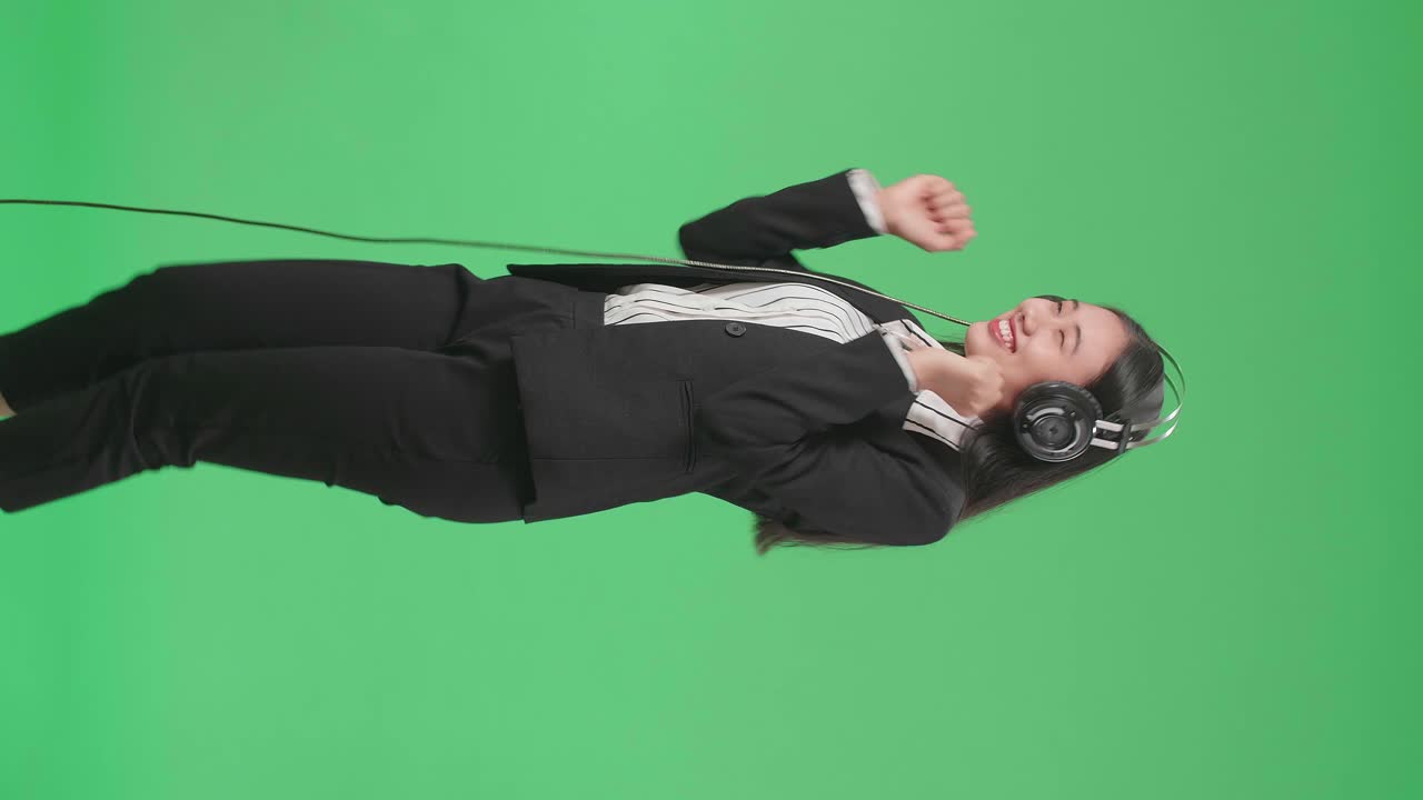 Side View Of An Asian Business Woman Listening To Music With Headphones And Dancing In The Green Screen Studio