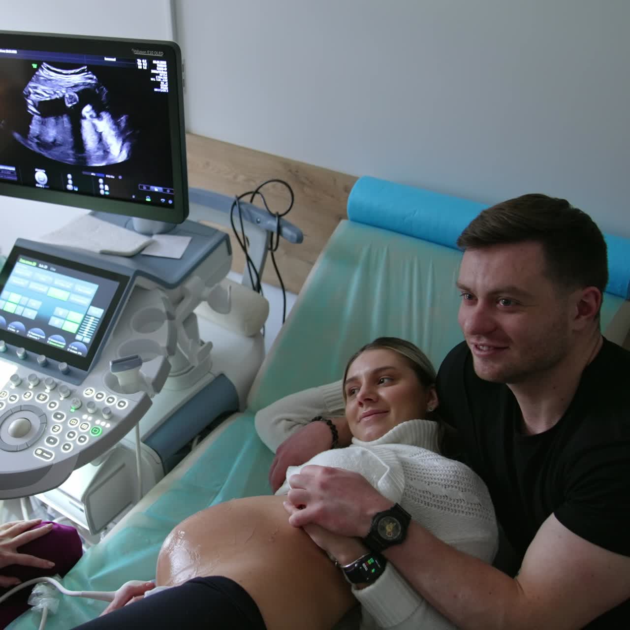 Happy smiling expecting couple having a baby ultrasound check. Pregnant lady lies on her back and husband supports her. Top view