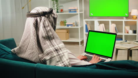 middle eastern man using laptop with chroma key close-up, typing on computer keyboard. television with green screen in living room. wearing traditional islamic male clothes
