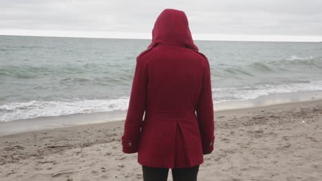 back view of a person wearing a red coat watching the sea waves while standing on the sandy shore - closeup shot