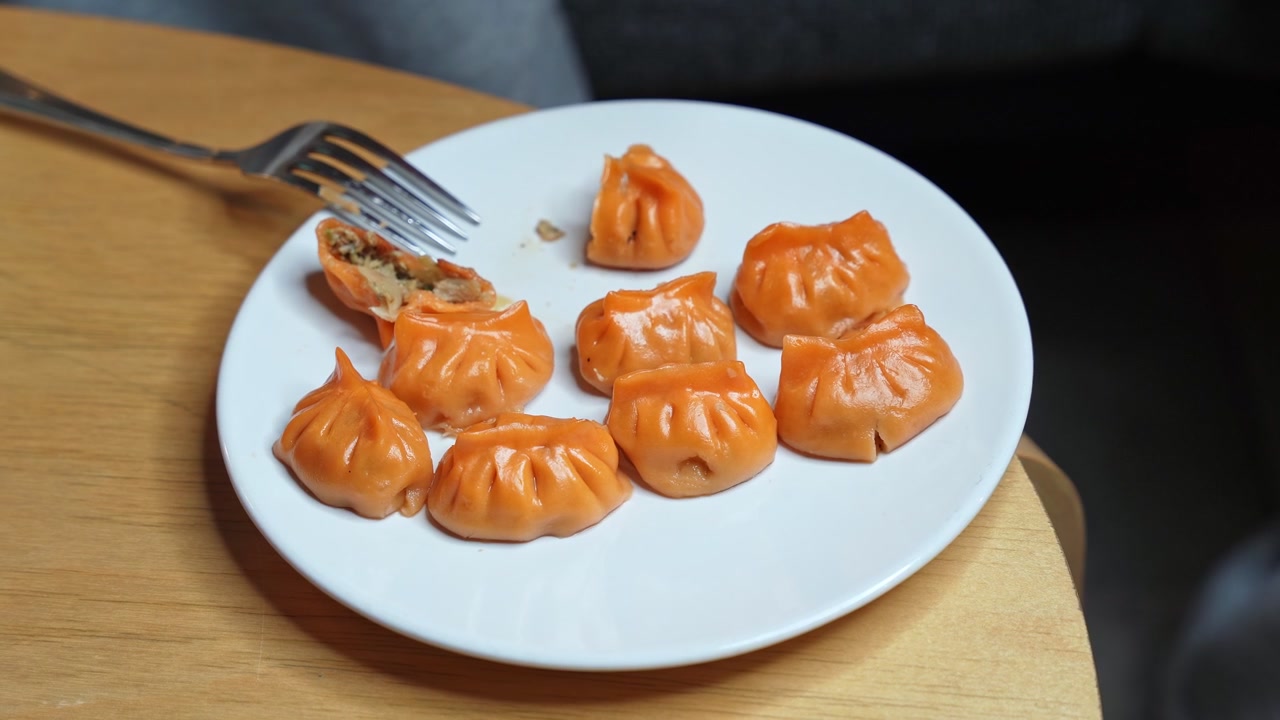 Foto close-up garpu perak menusuk pangsit Asia berwarna oranye untuk memperlihatkan isian gurih di dalamnya. Dim sum yang baru dimasak disajikan di piring putih yang diletakkan di atas meja kayu di dalam ruangan.