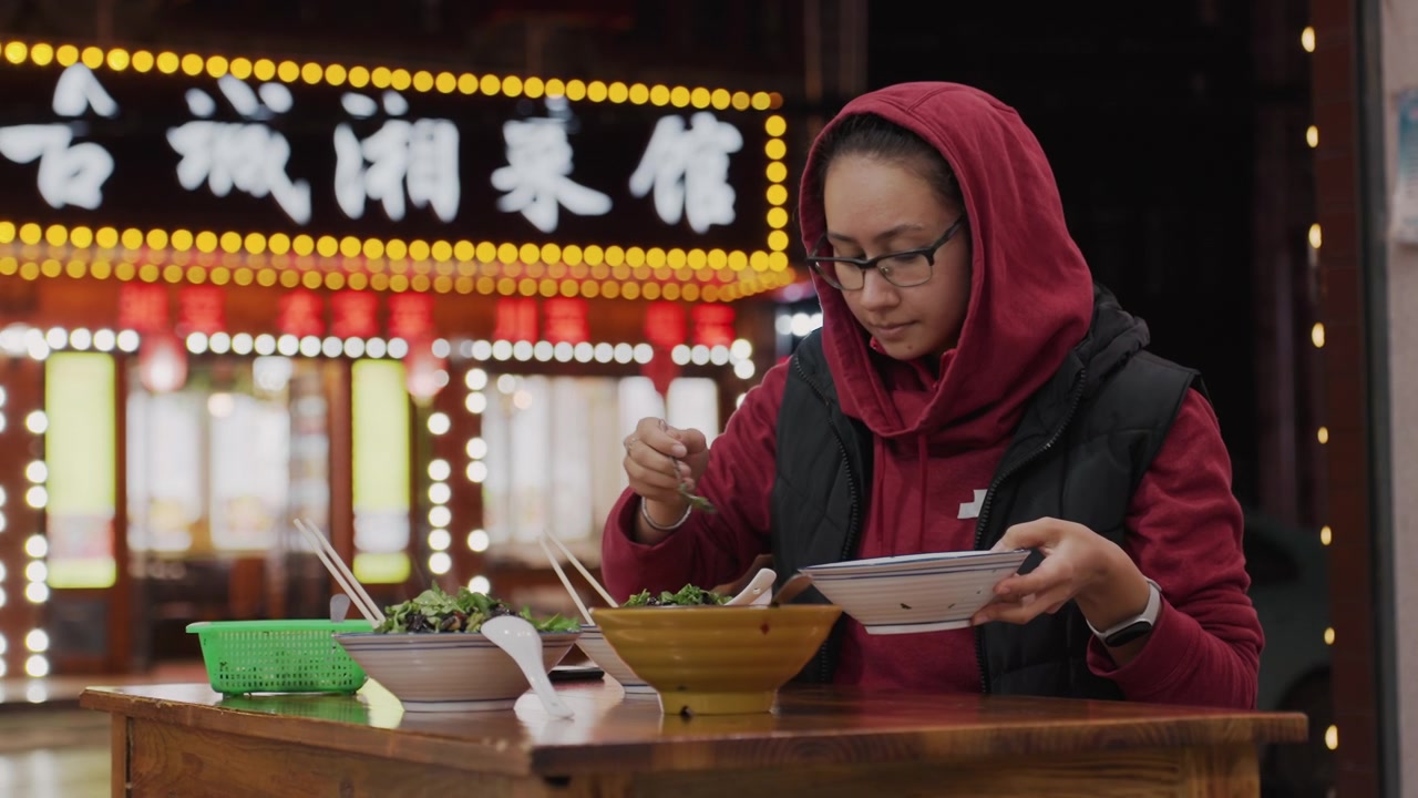 Night market scene woman eating noodles, neon signage glowing behind, steam rising from bowl, chopsticks in hands, scarf and hood framing face, glasses reflecting lights, wooden table with condiments