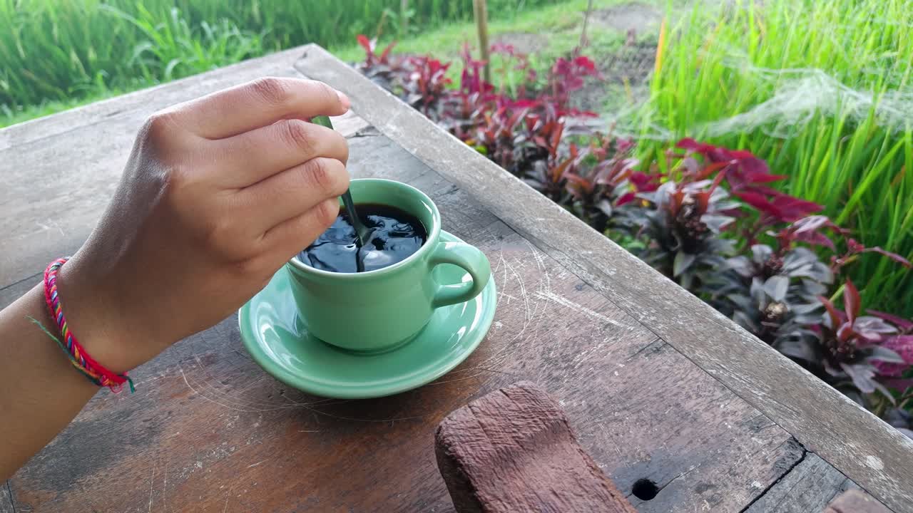 vista lateral mujer mano agitar negro luwak balí café con fondo de madera mesa arrozales archivados y vaso de taza verde sentirse feliz y energizado por la mañana