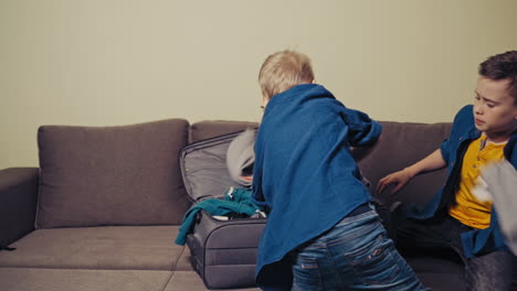 two curious boys are putting clothes into the suitcase for a trip at home. active brothers packing the things in the travelling bag together in the room. slow motion.