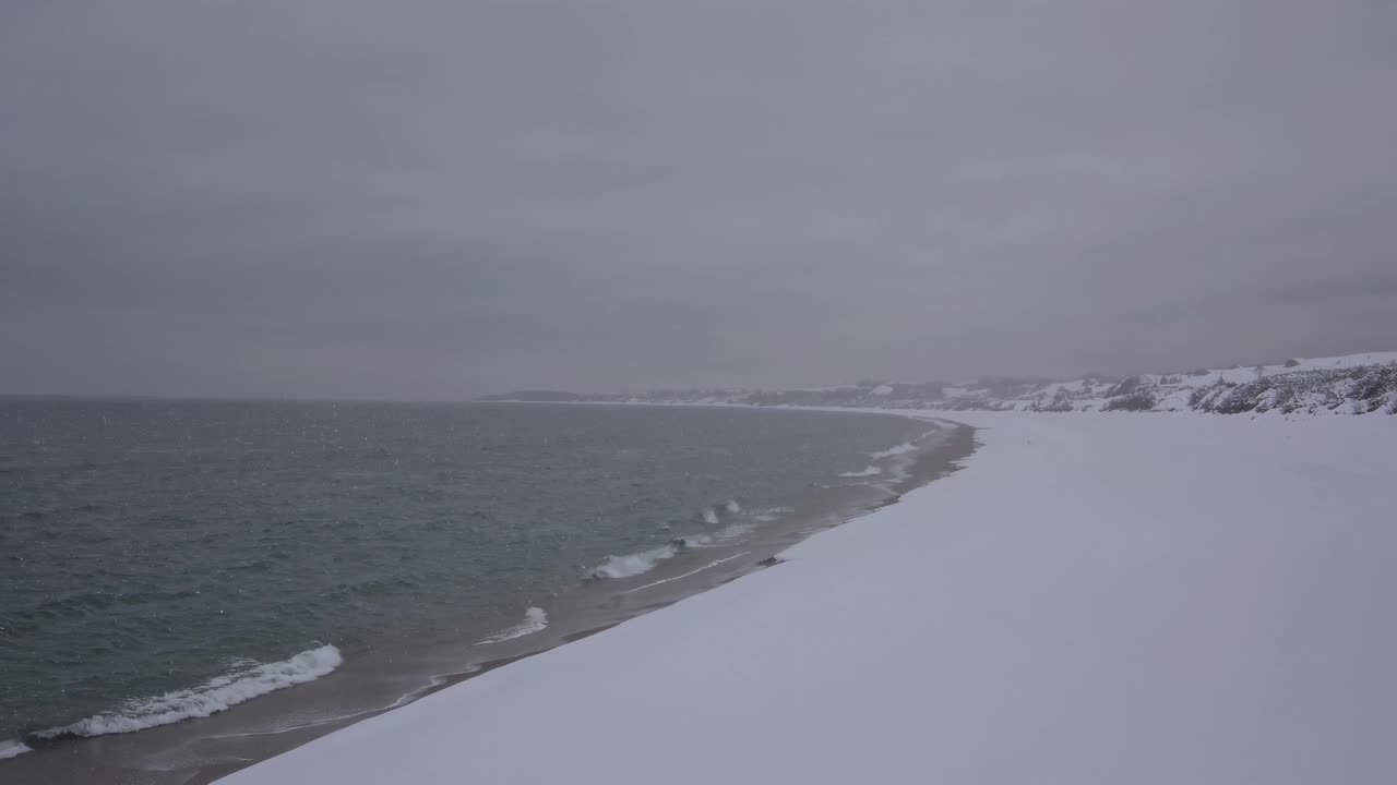 Aerial video captures a serene, snow-covered coastline meeting a calm sea under a gray sky