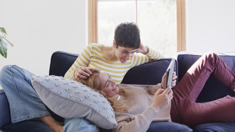 행복한 백인 레즈비언 커플이 소파에 누워 태블릿을 사용하고, 니 하우스에서 얘기하고 웃고 있습니다.