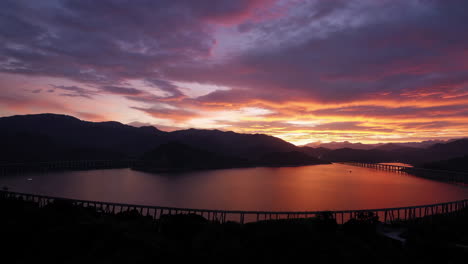 dramatic sunset over a lake with a long curving bridge and mountains