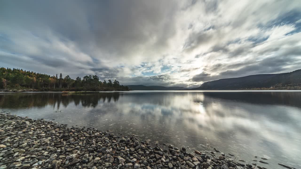 la superficie vidriosa del lago poco profundo con playa de guijarros refleja nubes blancas que se mueven rápidamente