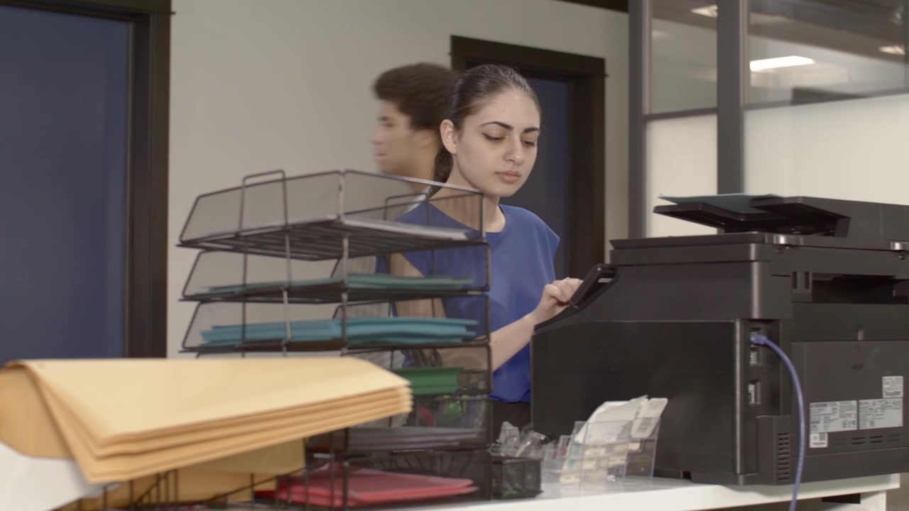 Mujer árabe-americana en una impresora en una oficina concurrida con movimiento de cámara de derecha a izquierda