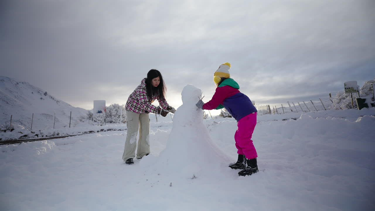 A mother and her young daughter happily build a snowman together in a wide, snowy landscape. This heartwarming clip is perfect for themes of family, winter fun, and childhood memories