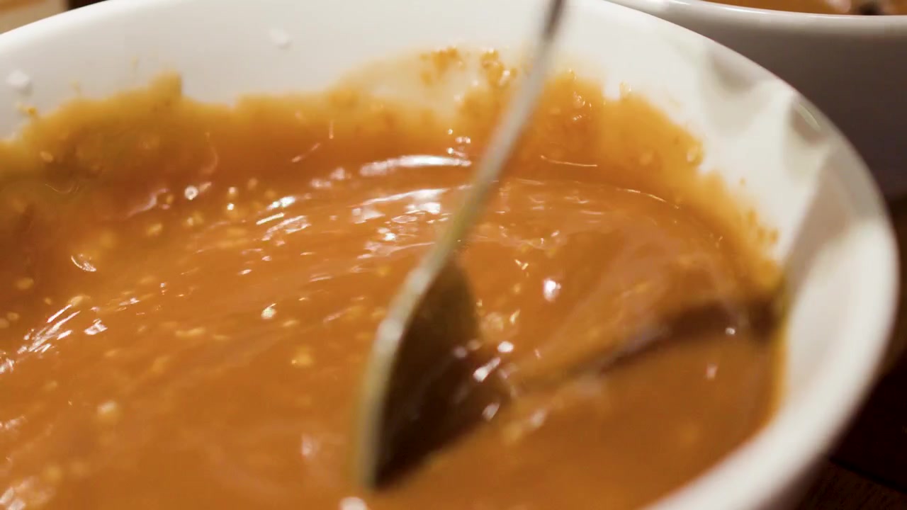 A metal spoon stirs thick brown sesame dipping sauce inside a white ceramic bowl