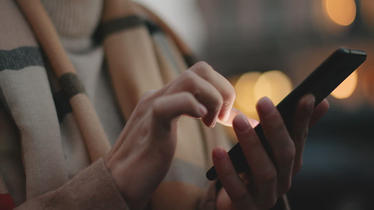 vista de primer plano de las manos de una mujer de negocios caucásica enviando mensajes de texto en su teléfono inteligente en la calle con luces de la ciudad en el fondo