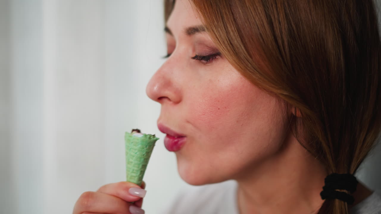 Close-up view of woman enjoying green ice cream cone with relaxed expression while standing near curtain indoors, soft light filters through sheer curtain