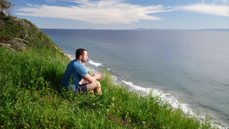 Un-Joven-Viajero-Sentado-Y-Disfrutando-De-La-Naturaleza-En-Un-Campo-De-Hierba-Al-Borde-De-Un-Acantilado-De-Playa-Con-Vistas-Al-Océano-De-Santa-Bárbara,-California-A-Cámara-Lenta