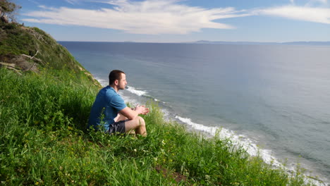 un joven viajero sentado y disfrutando de la naturaleza en un campo de hierba al borde de un acantilado de playa con vistas al océano de santa bárbara, california a cámara lenta