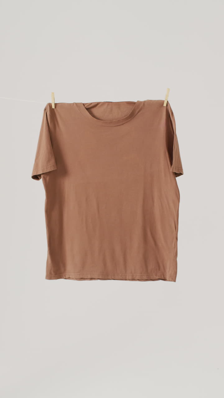 Vertical video of close up of brown t shirt hanging on white background with copy space