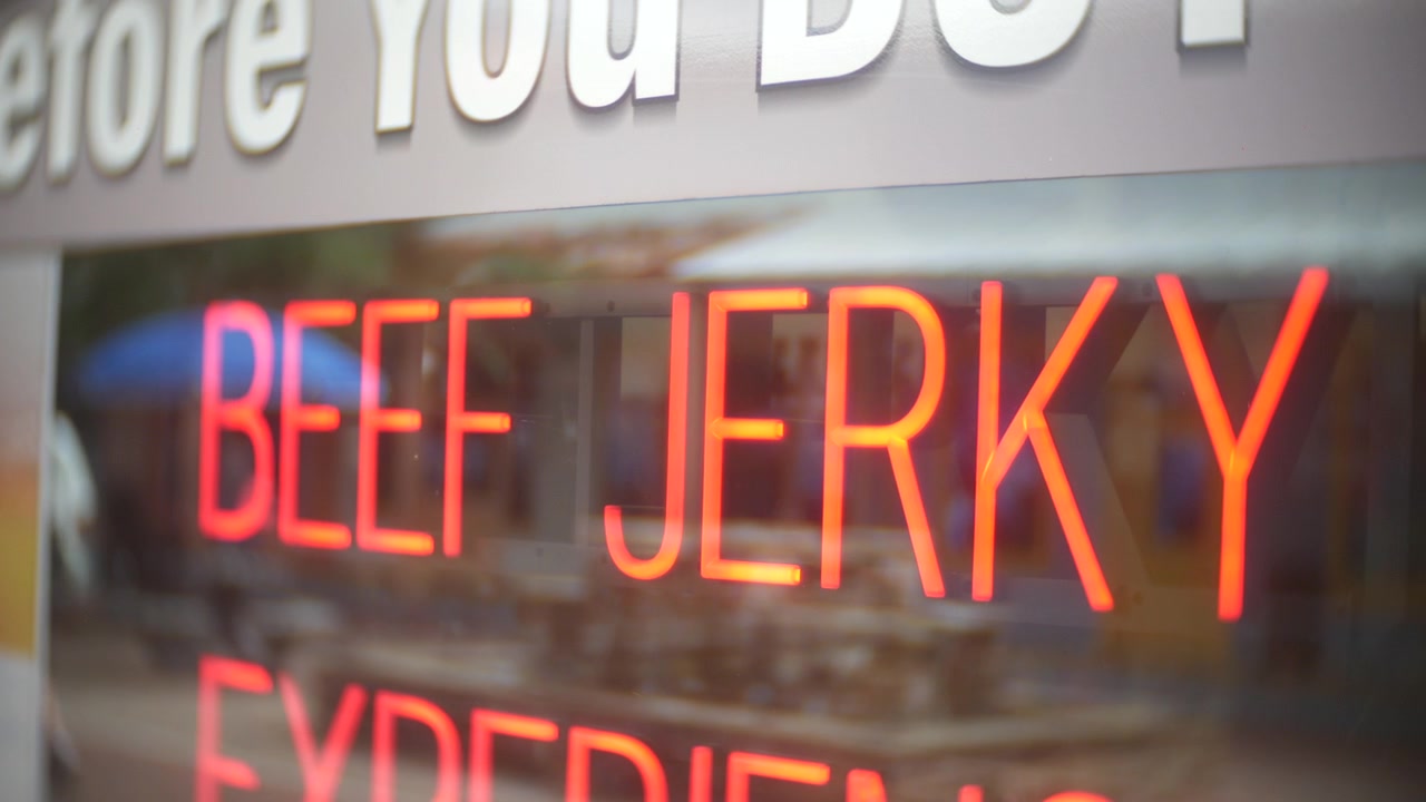 Insegna luminosa "Beef Jerky" in una vetrina con tenui riflessi. Una vista ravvicinata mostra scritte al neon rosse, l'interno sfocato del negozio e un espositore di alimenti al dettaglio.