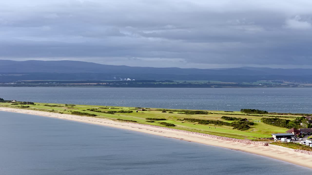 Drone camera slowly pans over Rosemarkie beachfront, caravans, and coastal landscape under cloudy daylight