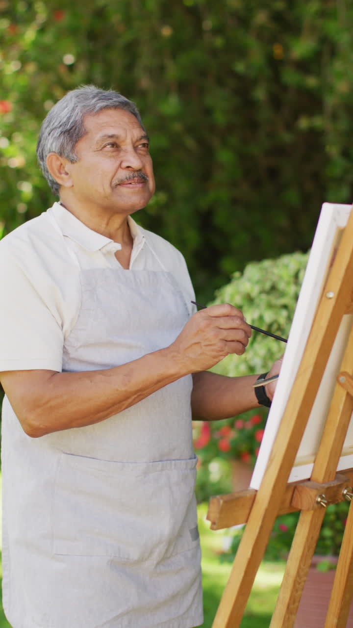 video vertical de un hombre bi-racial senior feliz pintando en un día soleado en el jardín