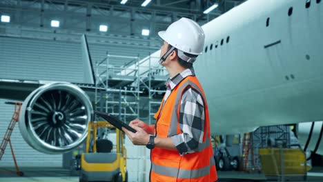 vista lateral de un ingeniero masculino asiático con casco de seguridad mirando la tableta en su mano y mirando a su alrededor mientras está de pie con aviones en el hangar