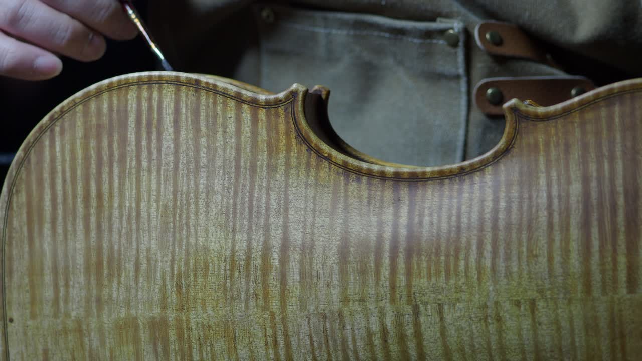 Skilled violinmaker carefully applies aging varnish along the ribs of a violin, revealing the marbled flames of the back plate in workshop setting, captured in close-up detail shot