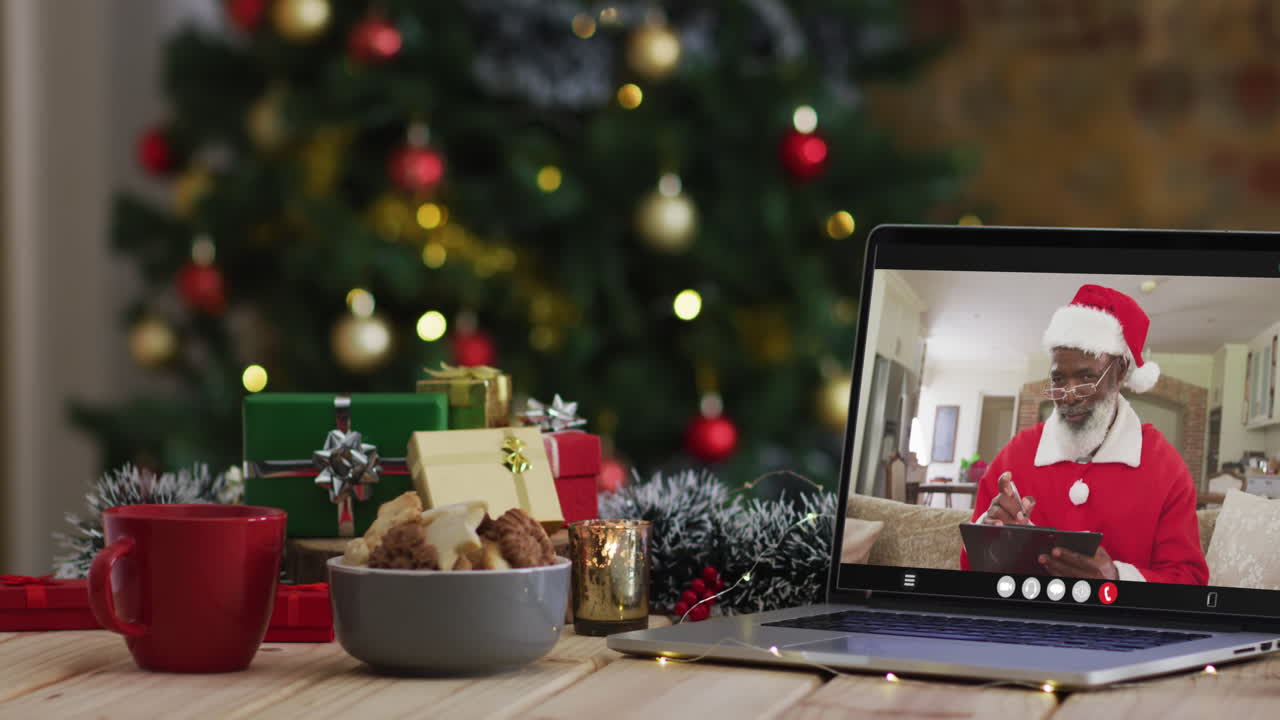 hombre afroamericano senior en disfraz de santa en videollamada en computadora portátil, con árbol de navidad