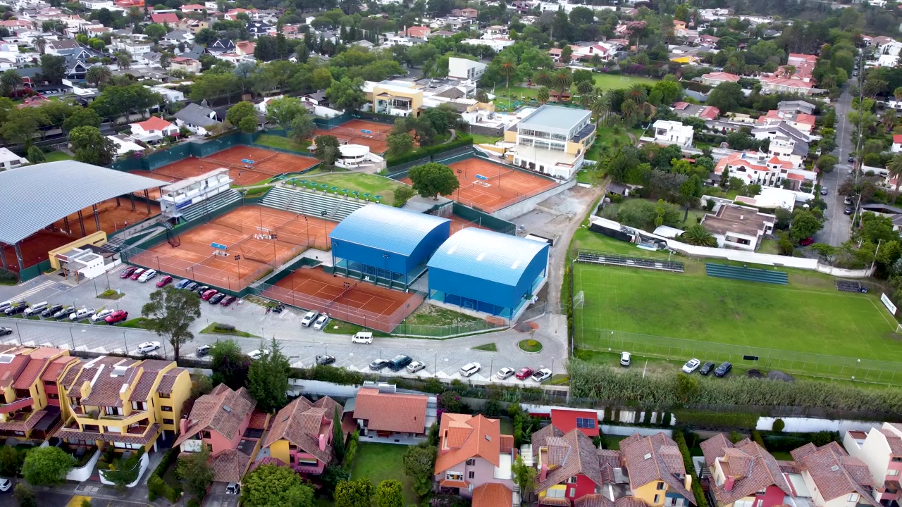 Uma vista aérea do complexo esportivo finalizado em Quito, Equador, apresentando os edifícios de telhado azul concluídos, quadras de tênis, campos de futebol e as áreas residenciais ao redor