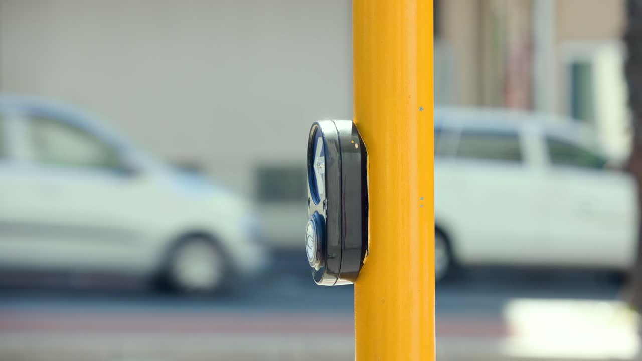 Person's left arm in maroon sleeve pressing walk button on bright yellow pole, needing to cross