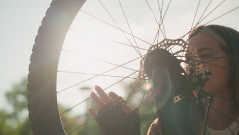 primer plano de un adulto joven tocando suavemente la rueda de la bicicleta mientras la luz del sol se refleja en ella, la rueda gira lentamente, y ella aparece ligeramente borrosa, en un fondo de vegetación vibrante