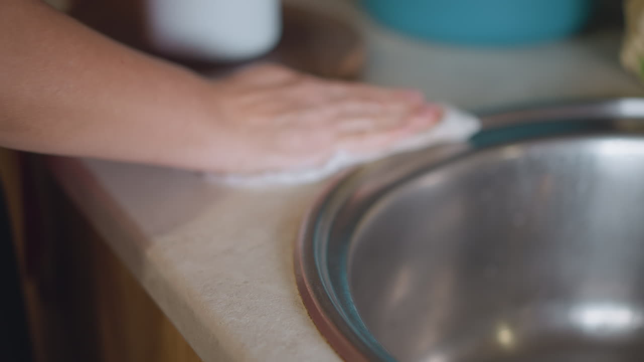 Mani adulte che puliscono il piano della cucina, cuoco casalingo che pulisce il bordo del lavello con un panno dopo aver cucinato, primo piano del movimento del panno sul lavello in acciaio inossidabile e sulla superficie in pietra, delicata faccenda domestica di routine con movimenti morbidi