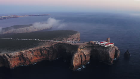 aerial shot of a lighthouse on rugged cliffs, surrounded by deep blue ocean and misty skies at purple sunset. sagres, algarve, portugal