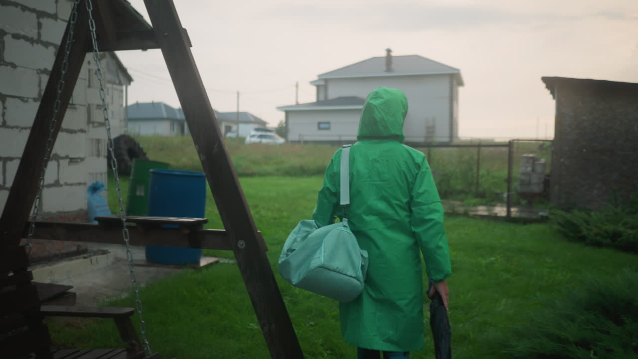 초록색 레인코트를 입고 민트색 가방과 우산을 들고 잔디을 향해 는 개인의 뒷면, 장면에는 스 벤치, 건물, 그리고 멀리 있는 자동차가 포함되어 있습니다.