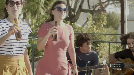 dos hombres sentados y tocando la guitarra mientras mujeres felices con gafas de sol bailando y bebiendo cerveza durante una fiesta en la azotea