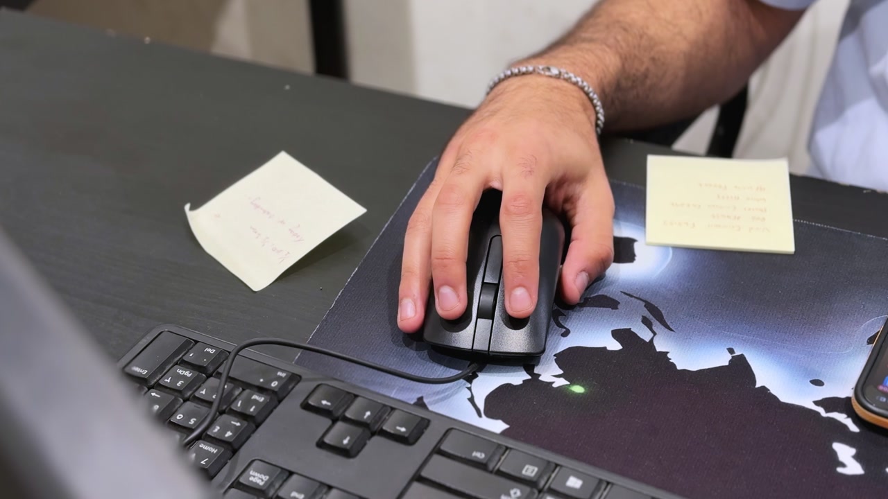 Close-up of a hand using a computer mouse on an office desk with sticky notes, smartphone, and monitor in an indoor workspace under soft lighting.