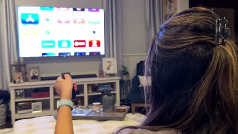 vista trasera de una mujer desplazando aplicaciones de transmisión en la televisión, usando el control remoto, en casa
