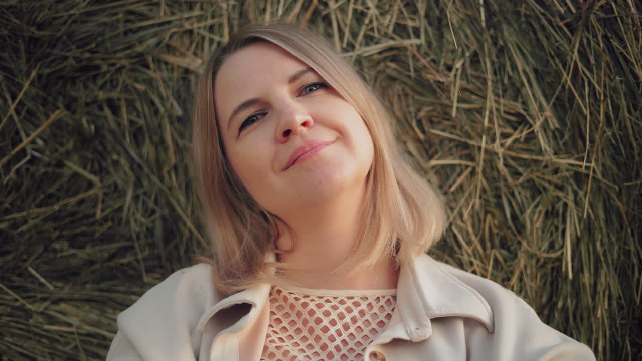 mujer con camiseta y chaqueta se apoya contra la bala de heno con una sonrisa suave y cálida, la luz del sol arroja un brillo suave en su rostro, creando un momento de paz y serenidad en un paisaje rural abierto