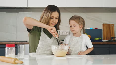 daughter-looks-at-mother-mixing-dough-with-whisk-at-table