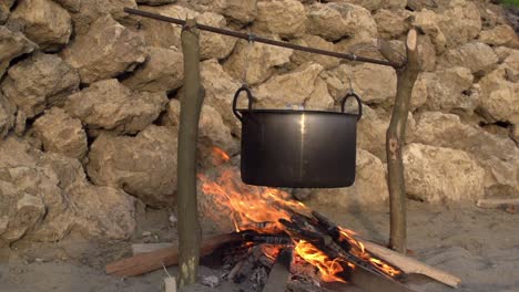 wide shot of a metal cooking pot hanging over an open fire on a sandy beach