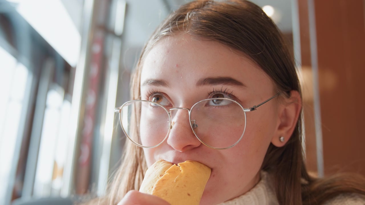Frontal close-up shows young woman in round glasses tasting wrapped sandwich inside warm cafe, with slight head tilt, calm gaze, soft daylight, and relaxed everyday food moment, with woman, glass