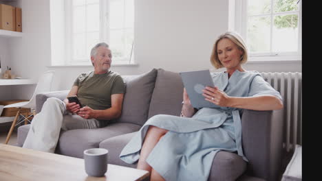 Senior-Couple-Sitting-On-Sofa-At-Home-Using-Digital-Tablet-And-Mobile-Phone-Together