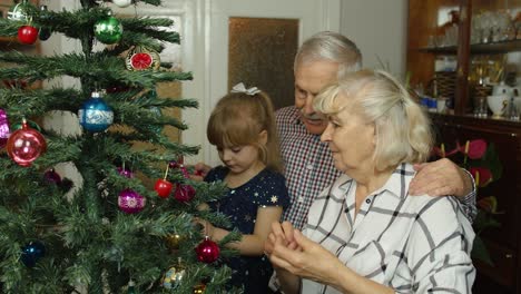 Girl-kid-with-senior-grandma,-grandpa-decorating-artificial-Christmas-tree-with-ornaments-and-toys